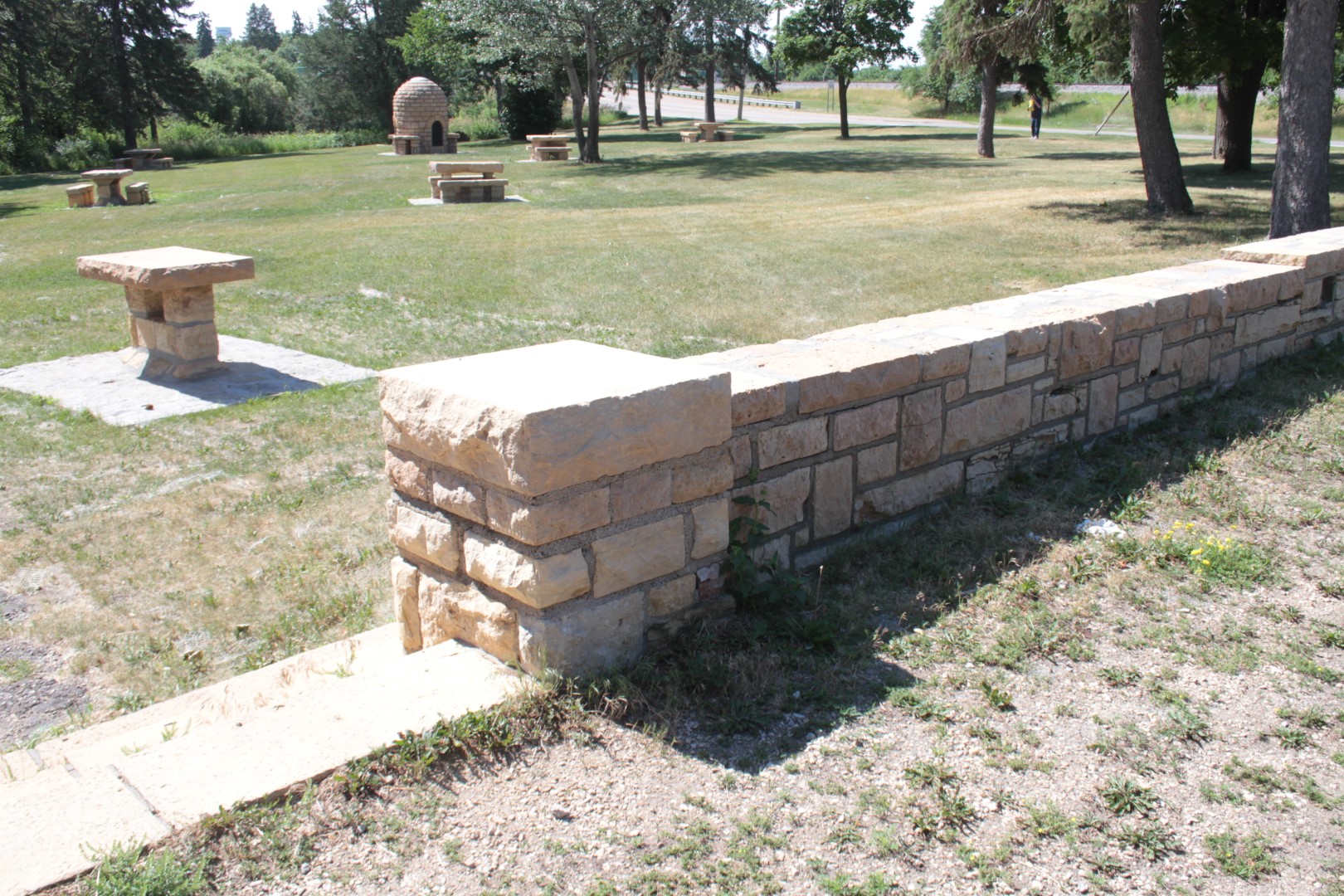 low stone wall with opening and a few steps into a grass picnic area with stone picnic tables and the “beehive” fireplace