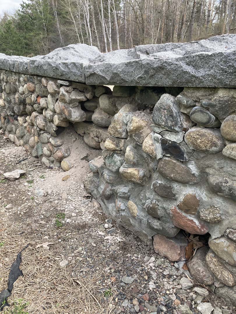 close-up of cobblestone retaining wall with crumbling mortar and stones missing and fallen out of wall. Long rectangular cap stones with rusting tops continue over damaged wall area. Light is visible through wall. Evergreen and light trunked trees in background across road