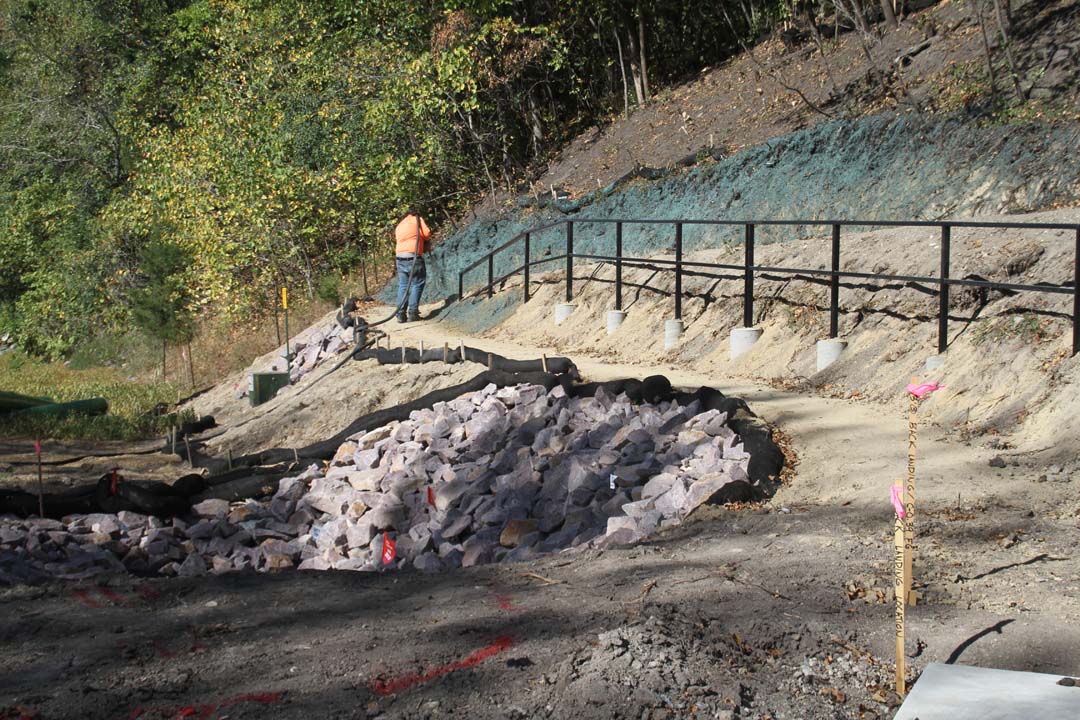 a workers standing on a dirt path spraying green “hydro seed” on a slope with a black metal rail separating two trail segments