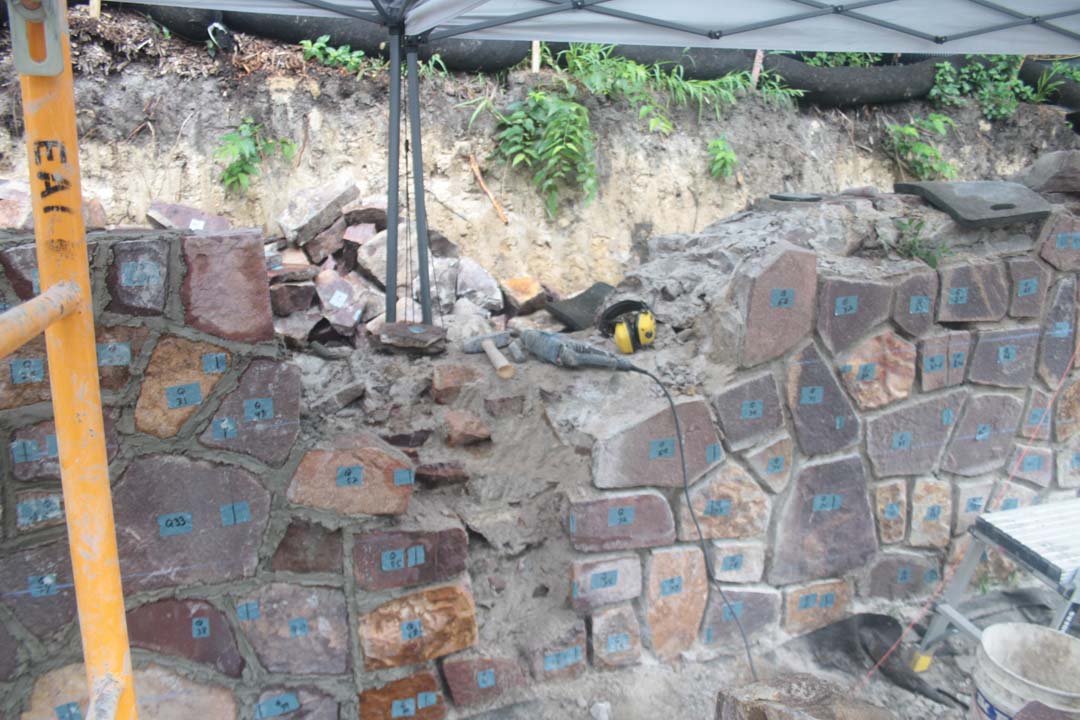 close view of a stone wall partly disassembled and scattered tools on top. Stones are irregularly shaped with flat faces and have tape with numbers on them