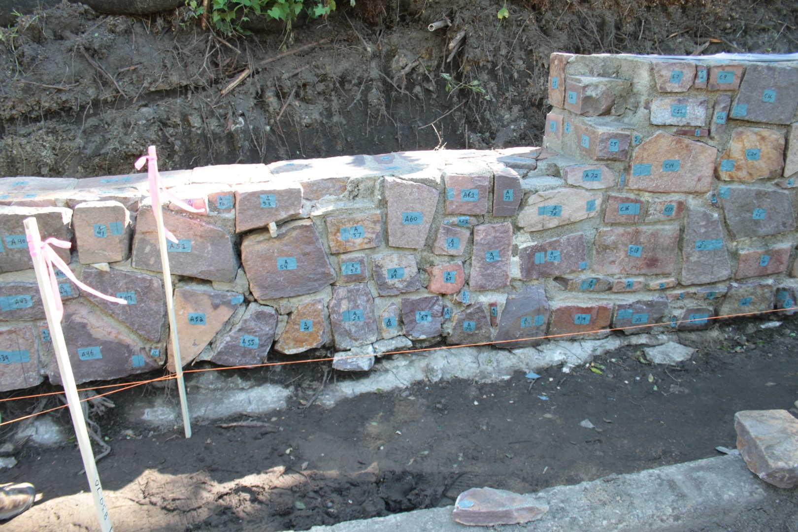 short stone wall with mortar removed stones have tape with numbers and exposed dirt below and in back of wall 