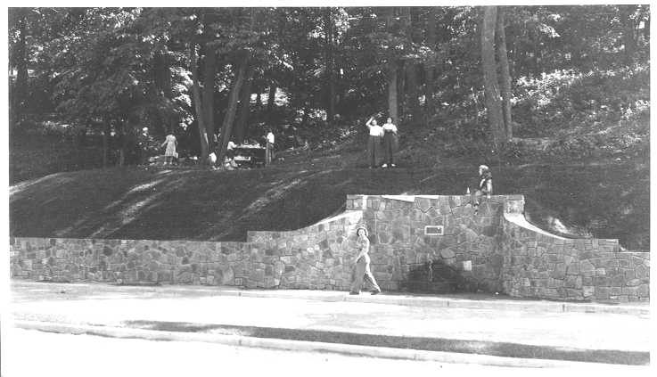 historic photo from 1939 of wayside adjacent to road: a tall stone retaining wall with a central spring pipe flowing with water and curved walls decreasing in height. Several women, one standing on walk below wall, one sitting on top of wall with legs dangling over, two people standing above wall on grass. A few more people visible in background at a picnic table. All people are women wearing 1940s clothes-dark jeans button shirts, only one is wearing a full skirt