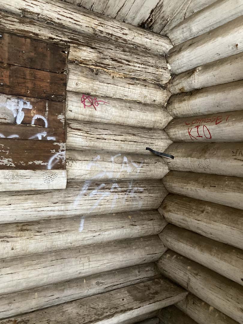 inside corner of log structure-round logs are whitewashed and closely fitted to each other. Dirt and graffiti on walls, window blocked with unpainted wood boards