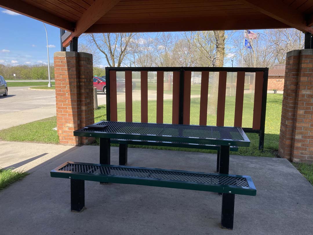 concrete walk at edge of parking area and brick piers and low sloping roofed picnic shelter with freestanding wind scree and shaded picnic tables inside. Trees in background